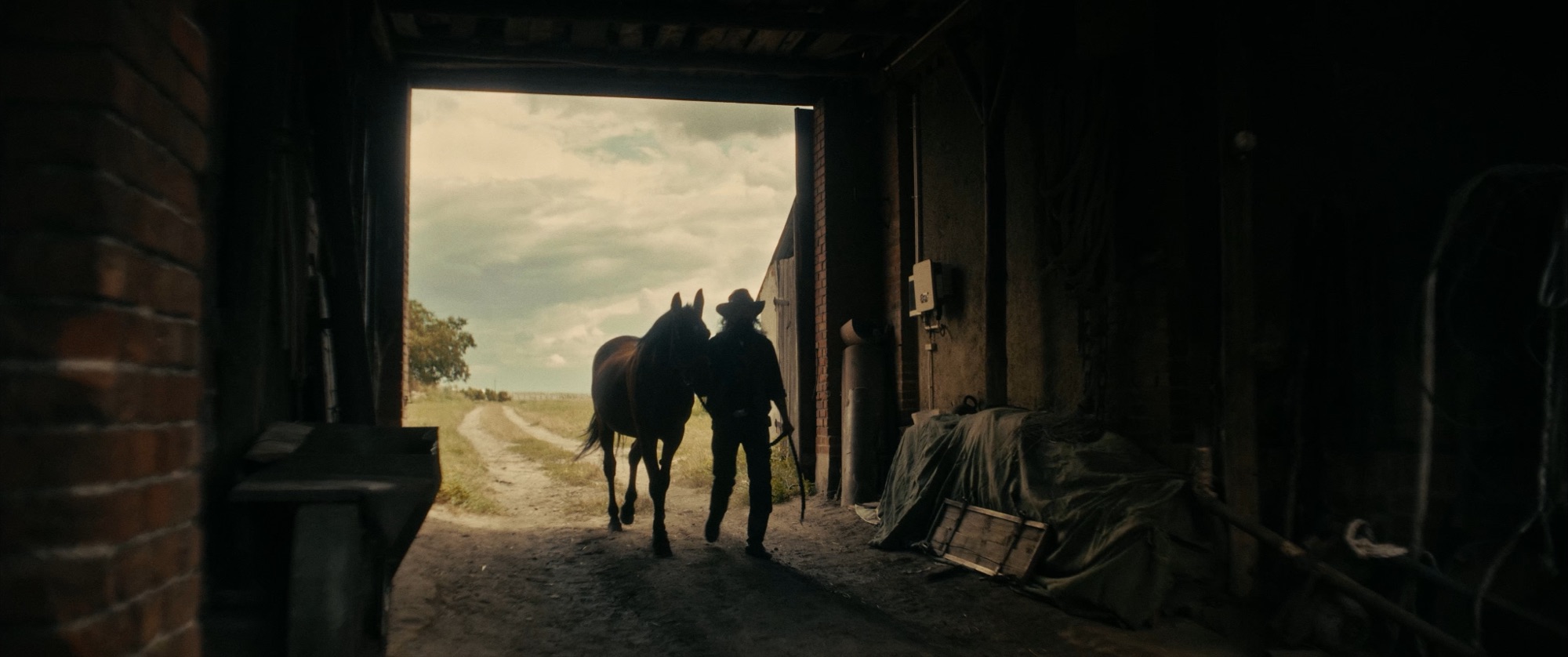 Silhouetted cowboy leading a horse out of a dark barn toward a sunlit field and cloudy sky - Amon Amarth